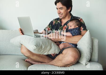 Father working on his laptop on the sofa and holding his baby. Stock Photo