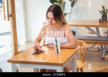 Focused young female freelancer in summer outfit working on smart phone while sitting in modern cafe Stock Photo