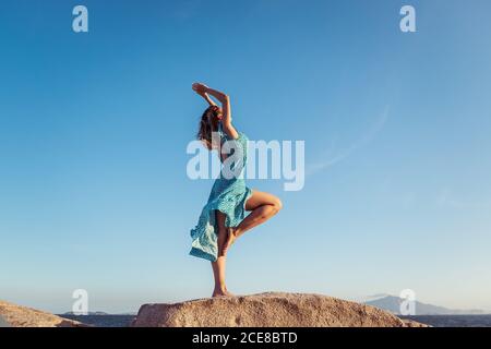 Side view of young happy slim female in stylish light dress posing on rocky cliff against cloudless blue sky during summer holidays Stock Photo