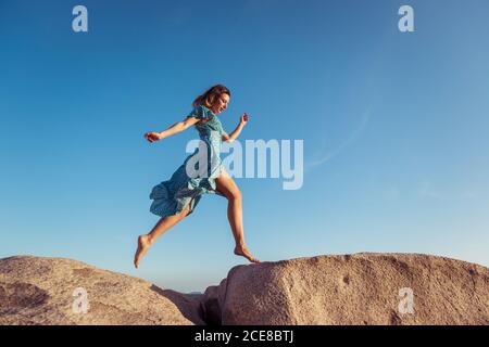 From below of young happy slim female in stylish light dress jumping on rocky cliff against cloudless blue sky during summer holidays Stock Photo