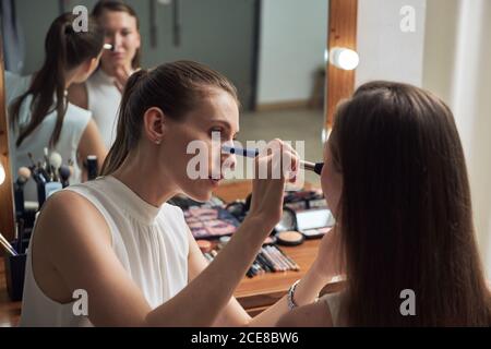 Positive young female makeup artist applying powder on clients face by using brush against cozy big mirror with lights in contemporary beauty salon Stock Photo