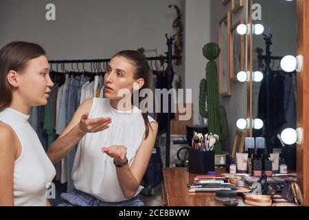 Concentrated female visagiste applying make up base with hands on customer sitting against cozy mirror with lights in modern studio Stock Photo