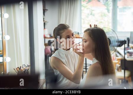 Positive young female makeup artist doing clients face makeup using brush near cozy big mirror with lights in contemporary beauty salon Stock Photo
