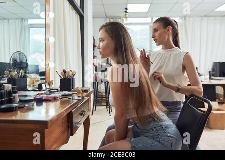 Positive young female makeup artist with brush standing near attractive client looking in mirror and checking makeup in modern studio Stock Photo