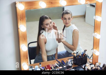 Positive young female makeup artist doing clients face makeup using brush looking at cozy big mirror with lights in contemporary beauty salon Stock Photo