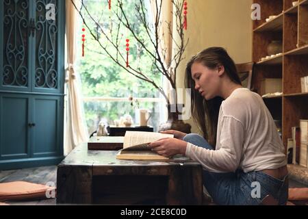 Side view of concentrated young female student in casual clothes reading textbook while sitting on floor at low table in oriental cafe Stock Photo