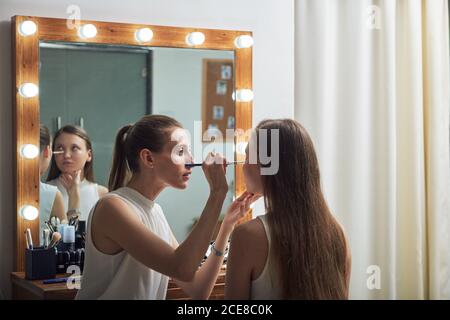 Positive young female makeup artist applying powder on clients face by using brush against cozy big mirror with lights in contemporary beauty salon Stock Photo