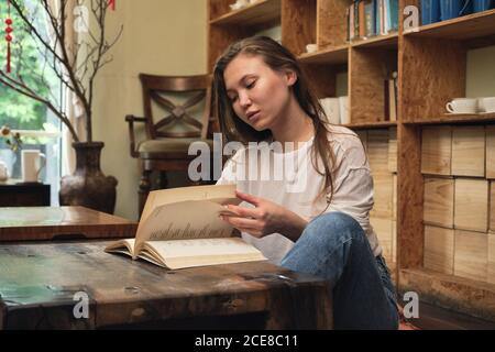 Concentrated young female student in casual clothes reading textbook while sitting on floor at low table in oriental cafe Stock Photo