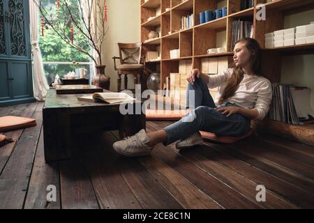 Side view of thoughtful young female student in casual clothes reading textbook while sitting on floor at low table in oriental cafe leaning on a bookcase Stock Photo