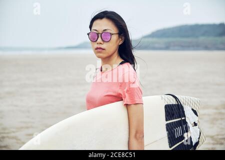 Young Asian female surfer in summer outfit walking on sandy beach and carrying surfboard against calm blue sea Stock Photo