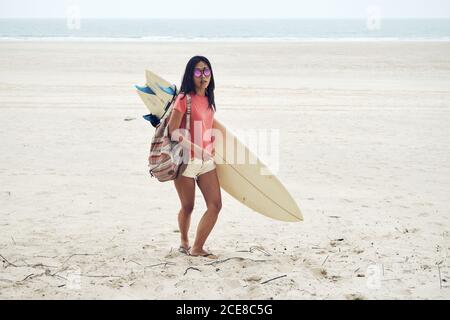Full length young Asian female surfer in summer outfit walking on sandy beach and carrying surfboard against calm blue sea Stock Photo