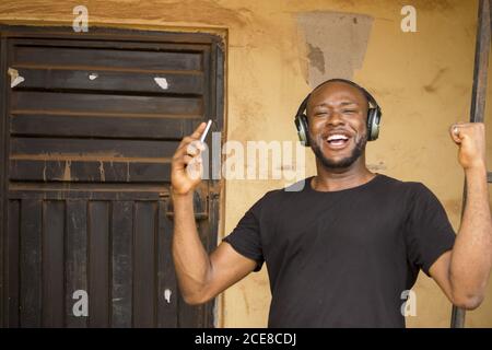 Young African male standing outside, using his wireless headphone to listen to music on the phone Stock Photo