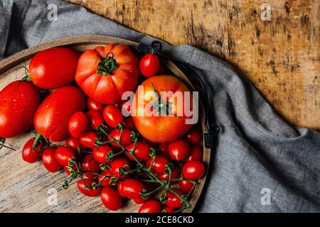 Top view of various types of fresh ripe red tomatoes on wooden tray arranged on rustic wooden table with cloth Stock Photo