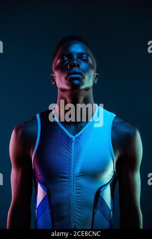 Serious concentrated young African American male athlete standing against blurred blue neon background in dark studio with eyes closed Stock Photo