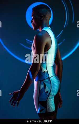 Side view of serious concentrated young African American male athlete standing against blurred blue neon background in dark studio looking away Stock Photo