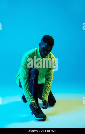 Full body serious young African American male athlete in bright colorful tracksuit tying shoelaces on sneakers in studio with blue neon illumination Stock Photo