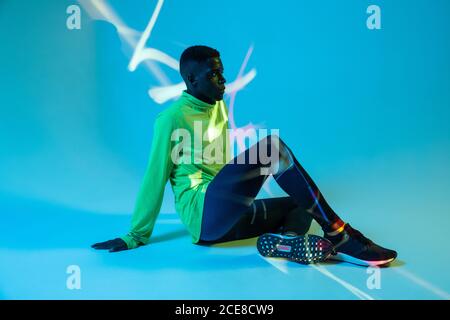 Full body side view of strong young African American male athlete in stylish colorful tracksuit and sneakers sitting on floor in blue studio with neon lights Stock Photo