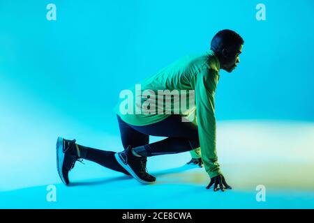 Full body side view of young African American male athlete in trendy sportswear and sneakers in low start position ready for sprint in studio with neon lights Stock Photo