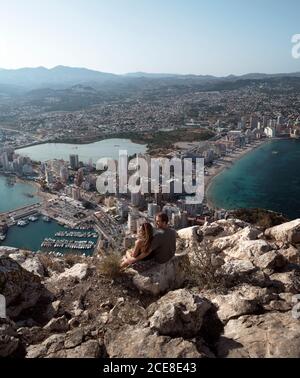 From above back view young couple embracing while observing fantastic view of Alicante while sitting on high rocky hill viewpoint during sunny summer day Stock Photo