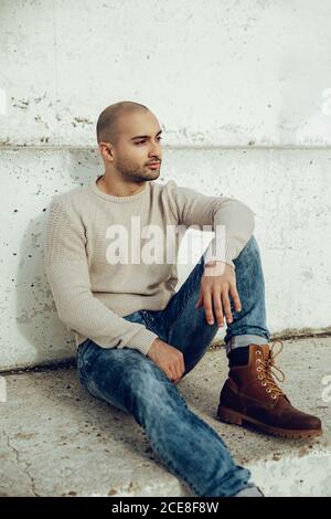 Pensive young unshaven guy in trendy sweatshirt and jeans with boots sitting on concrete stairs and looking away Stock Photo