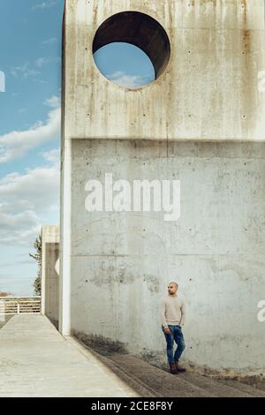 Full body young male in casual wear standing on stone stairs near weathered concrete construction and looking away Stock Photo