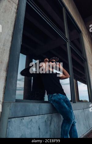 Low angle of confident young bearded male wearing black shirt and jeans with cap and sunglasses looking away while leaning against window of weathered stone building Stock Photo