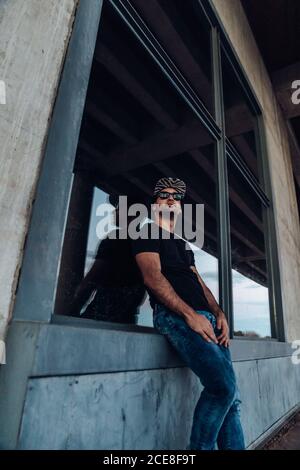 Low angle of confident young bearded male wearing black shirt and jeans with cap and sunglasses looking away while leaning against window of weathered stone building Stock Photo