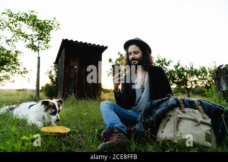 Delighted male hipster sitting on green filed in summer next to a dog and chatting on social media during weekend Stock Photo