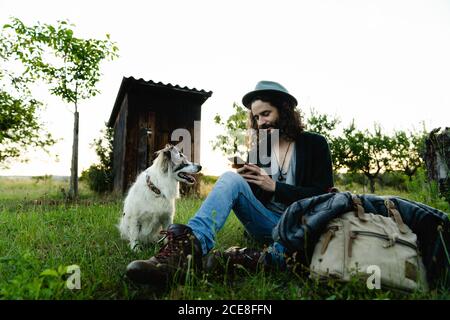 Delighted male hipster sitting on green filed in summer next to a dog and chatting on social media during weekend Stock Photo