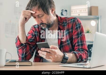 Troubled disappointed freelancer using smartphone for communication while sitting at the desk of his home office, selective focus Stock Photo