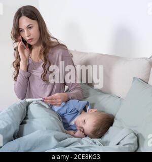 Worried young mother measuring temperature of her son and calling to a doctor. Sick kid feels bad and lying in bed under a blanket at home, focus on s Stock Photo