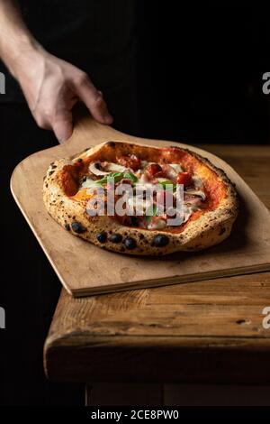 anonymous baker with delicious pizza with cheese and vegetables placed on wooden peel on black background Stock Photo