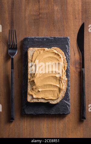 Top view of delicious piece of bread with peanut butter placed on black slate broad on wooden table with knife and fork Stock Photo