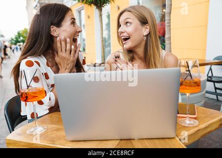 Image of delighted adult two women expressing surprise while using laptop at table in cafe outdoors Stock Photo