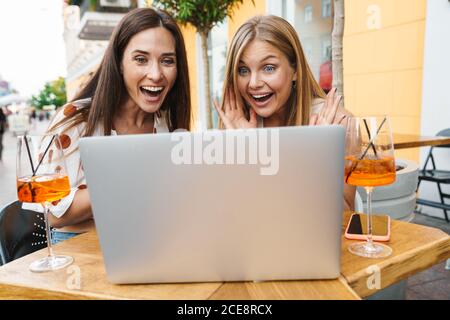 Image of delighted adult two women expressing surprise while using laptop at table in cafe outdoors Stock Photo