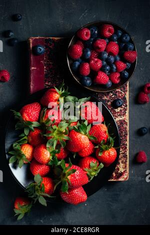 Top view composition with bowl with fresh ripe strawberries arranged near bowl with raspberries and blueberries on black surface Stock Photo