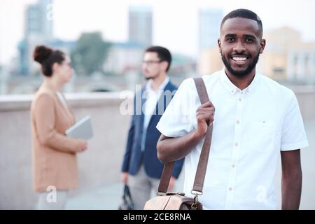 Portrait of cheerful Afro-American student guy with beard holding briefcase outdoors, their group mates talking in background Stock Photo