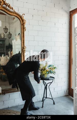 Side view of unrecognizable female designer in black outfit arranging floral composition on small table while working in creative studio with vintage interior Stock Photo
