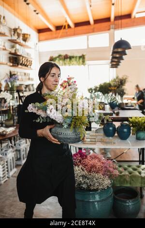 Professional female florist in black uniform carrying ceramic pot with bouquet of blooming flowers and green plants while working in floristry studio Stock Photo