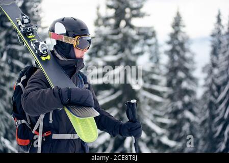 Portrait photo of male skier standing on slope top, looking at distance in his goggles. Professional sportsman in helmet with pair of skis. Grey snowy pine trees on blurred background. Monochrome view Stock Photo