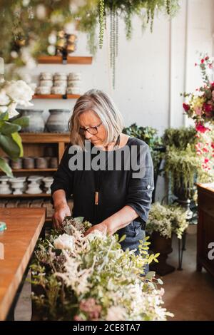 Professional adult female florist choosing green decorative plants for composition while working in cozy floristry studio Stock Photo