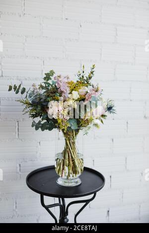 Beautiful bouquet with various flowers in ornamental glass pot placed on small table against white brick wall in creative floristry studio Stock Photo