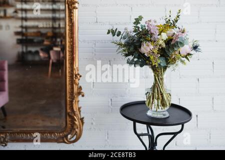 Beautiful spring bouquet with assorted colorful blooming flowers including pink peonies and blue sea holly flowers with mimosa twigs arranged on glass vase on table near white wall Stock Photo