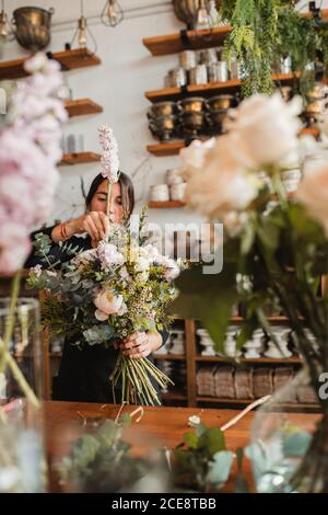 Concentrated young female floristry designer with big bouquet of fresh blooming flowers and green foliage standing at counter in cozy store Stock Photo