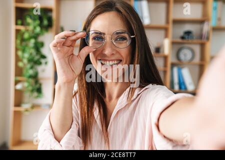 Image of joyful adult businesswoman smiling and holding eyeglasses while taking selfie photo in office Stock Photo