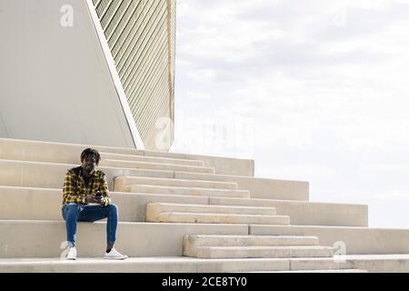 young black african-american man sitting on a concrete staircase with his phone, concept of lifestyle and technology, copy space for text Stock Photo