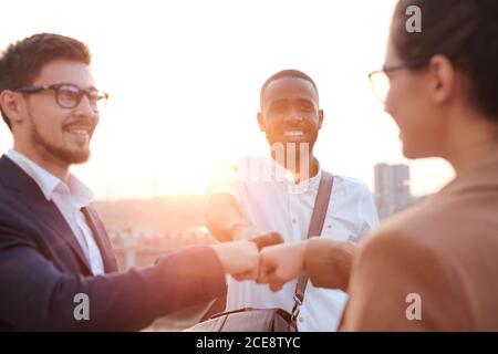 Group of positive young business colleagues making fist bump as symbol of teamwork in sunlight outdoors Stock Photo