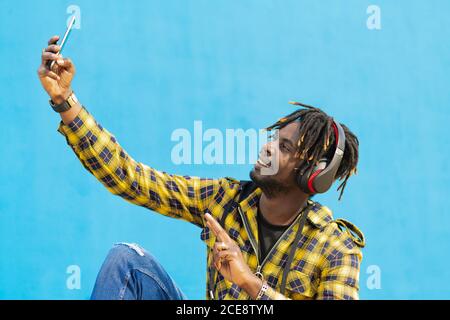 young black man with headphones taking a selfie with his smart phone with a blue wall in the background, technology and lifestyle concept, copy space for text Stock Photo