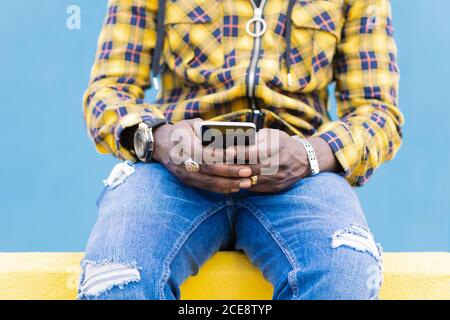 hands of an unrecognizable black man writing a message on his mobile phone sitting on a yellow wall with a blue wall in the background, technology and lifestyle concept, copy space for text Stock Photo