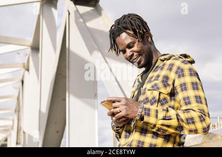 smiling young black man looking at his mobile phone with a modern metallic structure in the background, lifestyle concept, copy space for text Stock Photo